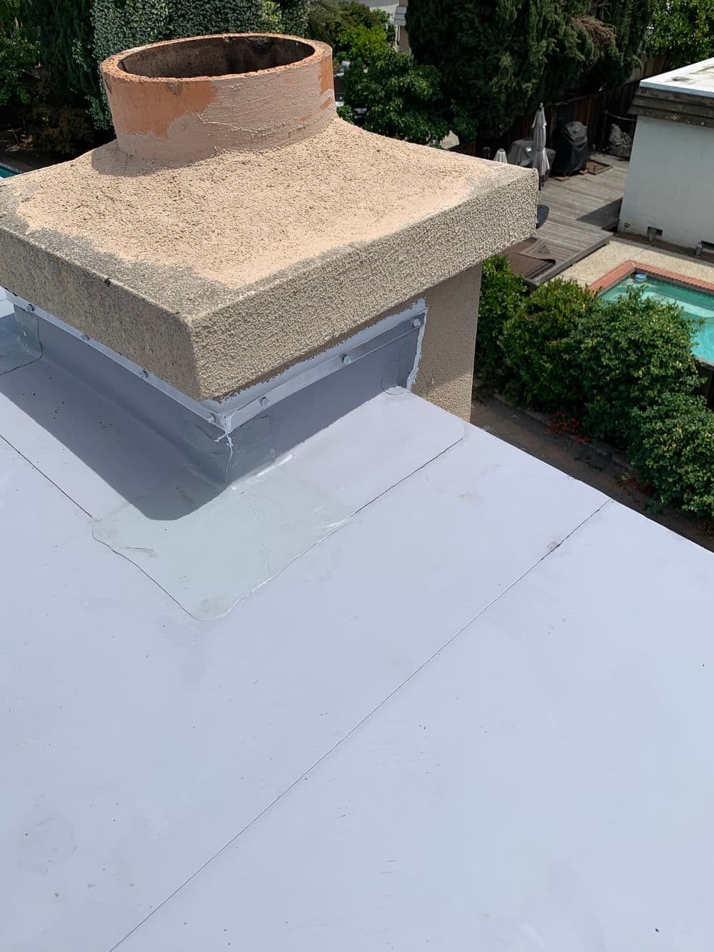 Roof with newly installed waterproof membrane and chimney, featuring green landscaping below.