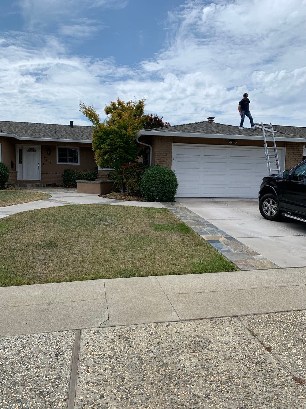 Person on a ladder cleaning the roof of a house with a well-manicured lawn and driveway.