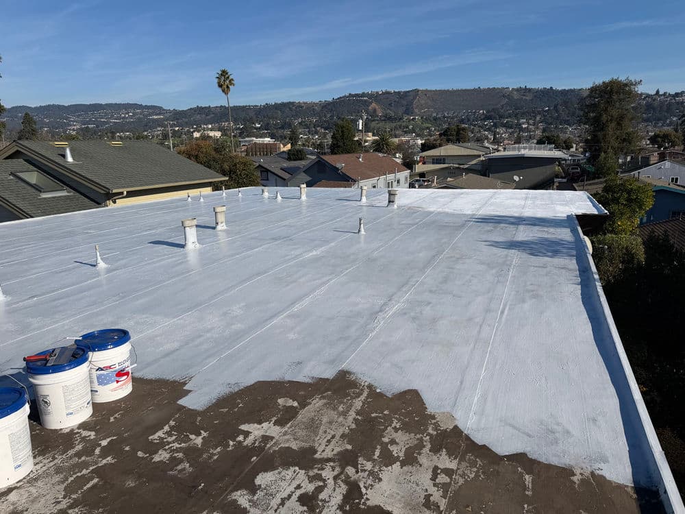 Flat roof coating application with white sealant, overlooking a residential area and hills.