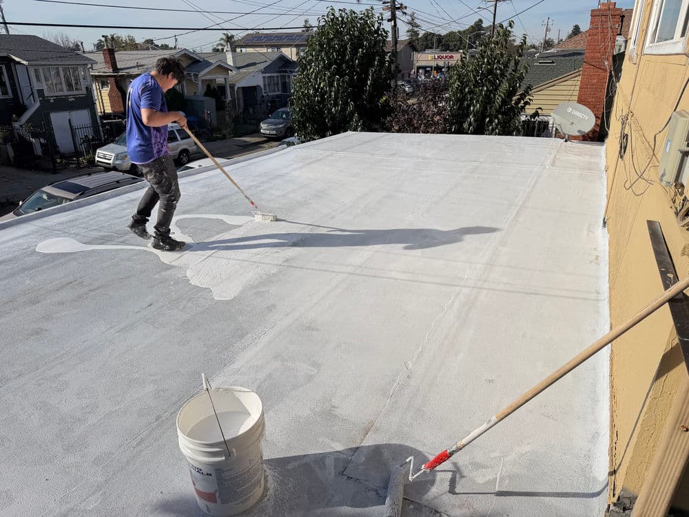 Man painting a flat roof with a roller, with a paint bucket nearby on a sunny day.